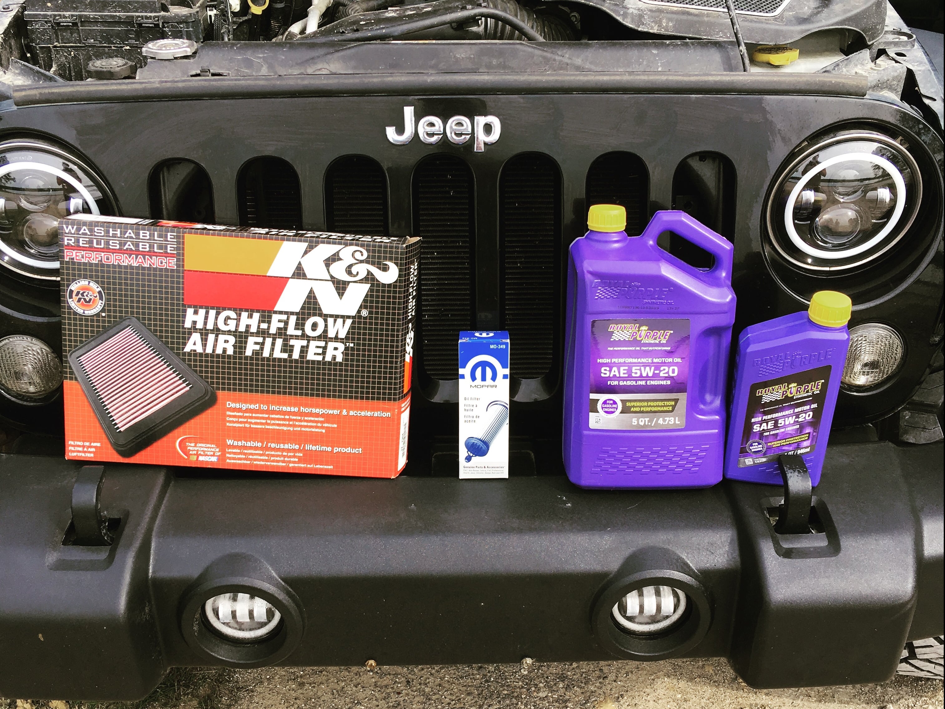 Engine maintenance supplies including synthetic motor oil, an oil filter, and a reusable high-flow air filter displayed in front of a Jeep Wrangler for a routine service.